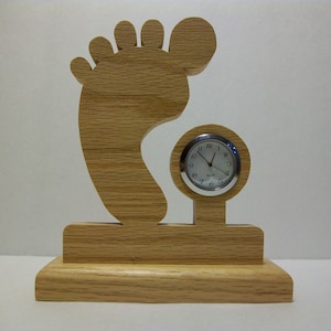 Puede incluir: Un reloj de madera con forma de huella de pie con una esfera redonda. La esfera del reloj tiene números y manecillas negras y un borde plateado.