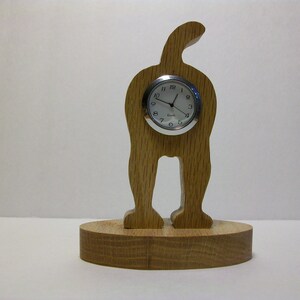Puede incluir: Un reloj de madera con forma de perro de pie sobre sus patas traseras. La esfera del reloj es redonda y plateada con números negros. El perro está hecho de madera marrón claro y tiene una cola que se curva sobre su espalda.