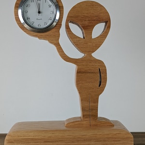 May include: A wooden alien figurine holding a clock with a white face and black hands. The clock face says "Quartz". The alien is standing on a wooden base.