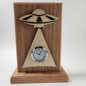 May include: A wooden clock with a UFO design. The clock face is white with black numbers and hands. The clock is on a wooden base.