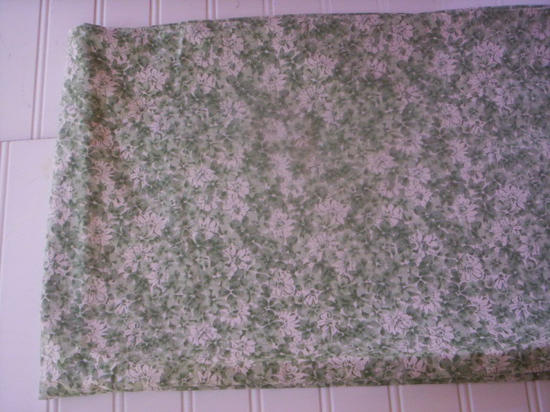 May include: A white fabric with a green floral pattern. The fabric is folded in half and has a smooth texture.