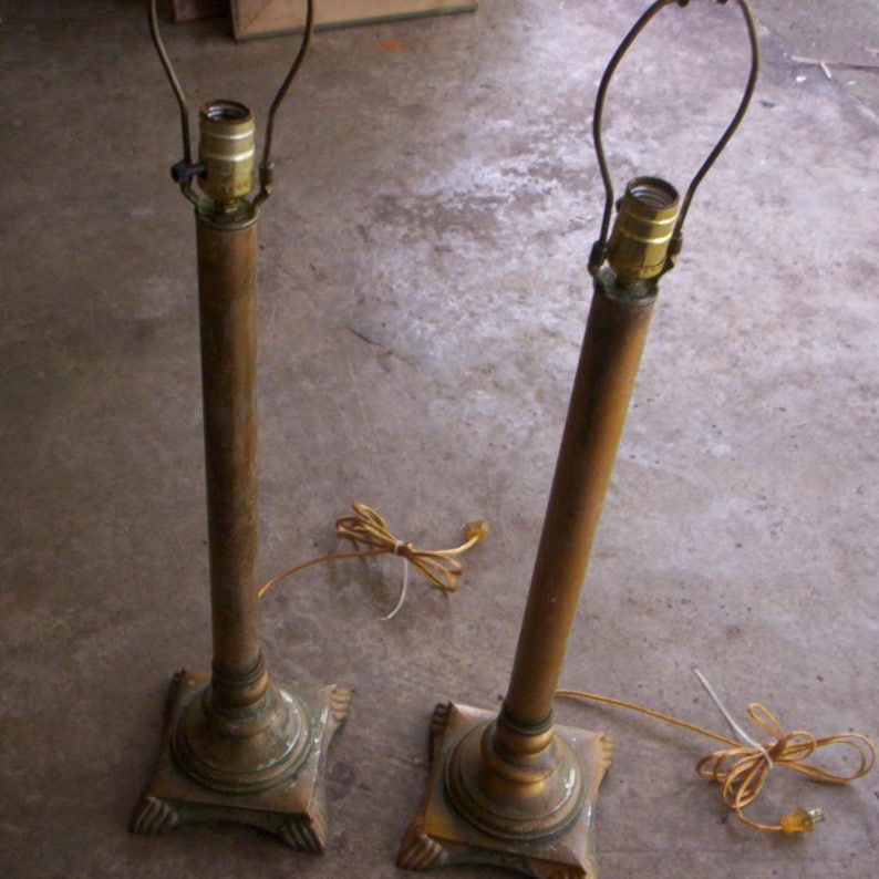 May include: Two antique brass floor lamps with a simple design. The lamps have a square base and a tall, slender column. The lampshades are missing.