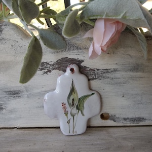 May include: White ceramic cross with a painted image of a white calla lily with green leaves. The cross is on a whitewashed wooden surface with a distressed finish.