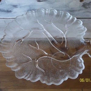 Puede incluir: Un plato de servir de vidrio transparente con forma de &aacute;rbol y superficie texturizada. El plato tiene un borde elevado y una peque&ntilde;a manija en el lateral.