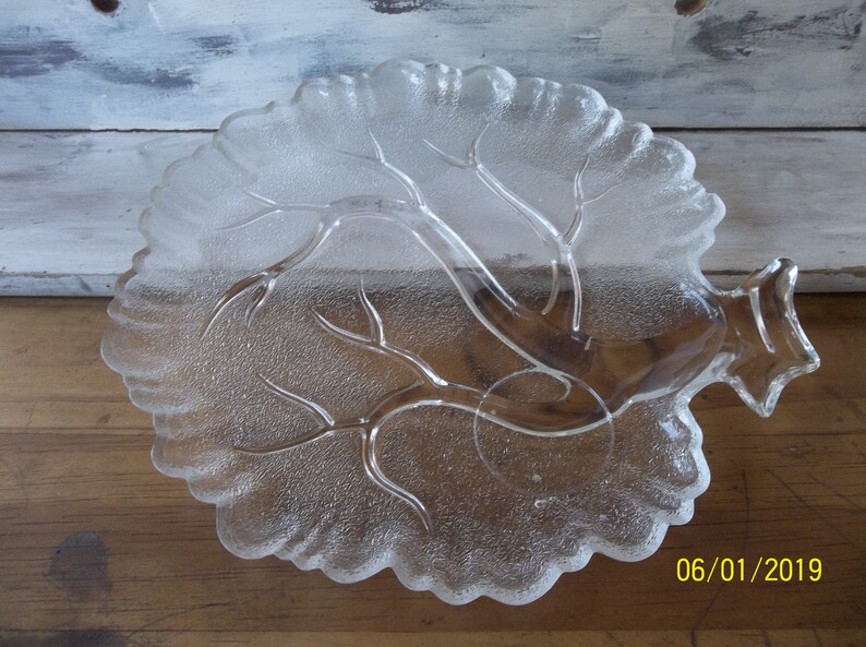 Puede incluir: Un plato de servir de vidrio transparente con forma de &aacute;rbol y superficie texturizada. El plato tiene un borde elevado y una peque&ntilde;a manija en el lateral.