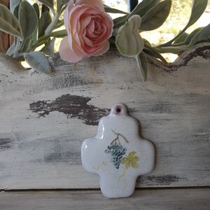 May include: A white ceramic cross with a painted design of a grapevine and grapes. The cross is hanging on a white wooden surface with a distressed finish.