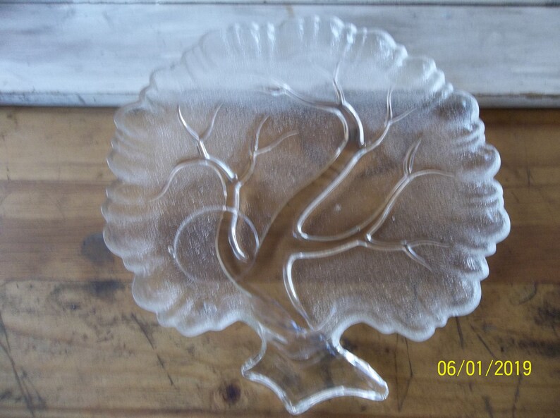Puede incluir: Un plato de servir de vidrio transparente con forma de &aacute;rbol con un dise&ntilde;o esmerilado. El plato tiene una base ancha y un tronco de &aacute;rbol que se ramifica en un dise&ntilde;o de hojas.