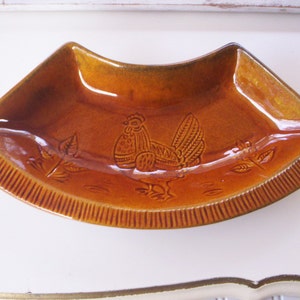 May include: A brown ceramic serving dish with a raised design of a rooster and leaves. The dish is shaped like a half-circle and has a scalloped edge.