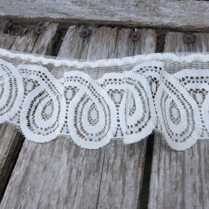 May include: White lace trim with a scalloped edge and a paisley pattern. The lace is on a weathered wooden surface.