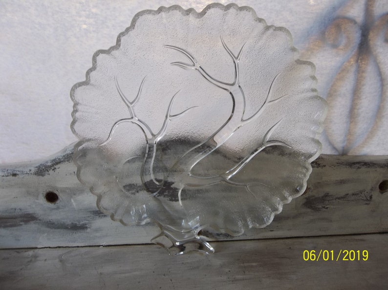 Puede incluir: Plato de servir de vidrio transparente con un dise&ntilde;o de &aacute;rbol. El plato tiene un borde festoneado y una base elevada.