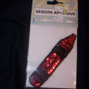 May include: Red sequin applique shaped like a crayon. The applique is 1 1/2 inches by 4 inches and is labeled "Sequin Applique" and "FSA301 A 1 1/2" x 4" GLOS-RED 1PC".
