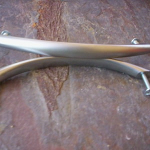Può includere: Due maniglie per mobili in nichel spazzolato con design curvo. Ogni maniglia ha viti per il montaggio. Le maniglie sono di colore argento e sembrano essere in metallo.