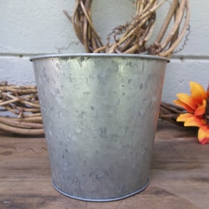 Puede incluir: Un macetero de metal galvanizado plateado con una superficie texturizada. El macetero está vacío y listo para plantar.