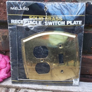 Pode incluir: Uma embalagem contendo uma placa de tomada e interruptor de latão maciço. A placa é dourada e tem uma borda arredondada. A embalagem é etiquetada como "Melard Solid Brass Receptacle/Switch Plate".