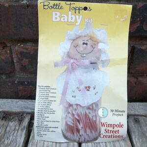 Può includere: Un kit per la creazione di una bambola per tappo di bottiglia. Il kit include una testa di bambola in feltro bianco, un nastro rosa, pioli di legno e un barattolo di bastoncini di cotone. La confezione include il testo "Bottle Toppos Baby Kit" e "30 Minute Project Wimpole Street Creations".
