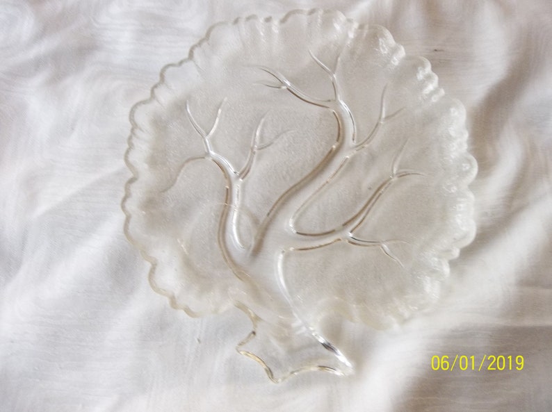 Puede incluir: Un plato de servir de vidrio transparente con forma de &aacute;rbol y borde festoneado. El plato tiene un dise&ntilde;o en relieve de ramas y hojas.