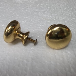 Könnte beinhalten: Zwei goldfarbene Schrankknöpfe mit runder, leicht gewölbter Form. Ein Knopf hat eine Schraube befestigt.