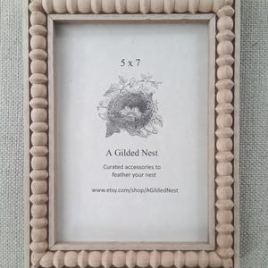 Op de afbeelding: Een lichtgekleurde houten fotolijst met een kralenrand. De lijst bevat een print met een nestillustratie en de tekst "5 x 7". De tekst "A Gilded Nest" en het websiteadres zijn ook zichtbaar.