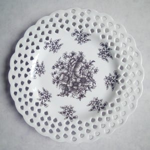 May include: A round white ceramic plate featuring a decorative lattice-style openwork border with a scalloped edge. The center of the plate is decorated with a detailed black floral illustration, surrounded by smaller matching floral motifs.