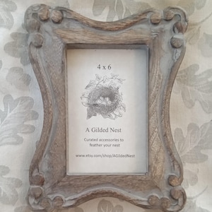May include: A distressed, gray-washed wooden picture frame with ornate detailing. The frame holds a 4x6 inch print featuring a nest illustration and the text "A Gilded Nest". The frame has a curved top and bottom.