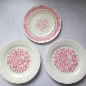 May include: Three ceramic plates featuring pink transferware patterns. The top plate has a floral center and a dotted border. The two bottom plates feature a central fruit basket design with grapes and peaches, surrounded by embossed rims.