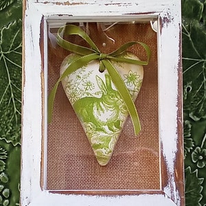 May include: A white wooden frame with a distressed finish, containing a green heart-shaped ceramic ornament with a rabbit and floral design. The ornament is tied with a green ribbon.
