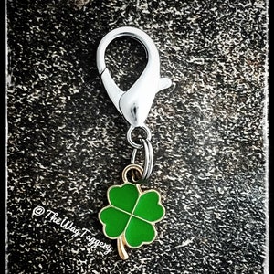 May include: A silver key chain with a green four-leaf clover charm. The charm is attached to a silver ring and a silver spring clasp. The background is a dark gray textured surface. @TheWagTaggery