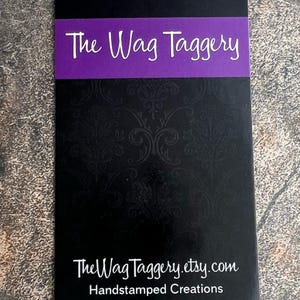 May include: Black business card with a purple banner that reads "The Wag Taggery". The text "TheWagTaggery.etsy.com" and "Handstamped Creations" are printed in white below the banner.