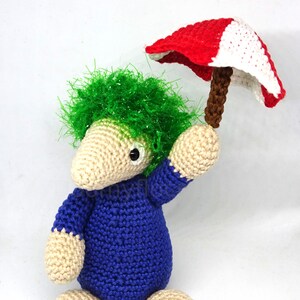 May include: A crocheted green-haired creature wearing a blue jumpsuit holds a red and white crocheted umbrella.