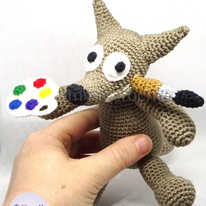 Puede incluir: Un lobo de crochet gris que sostiene una paleta de pintura blanca con puntos de pintura de colores y un pincel en la boca.