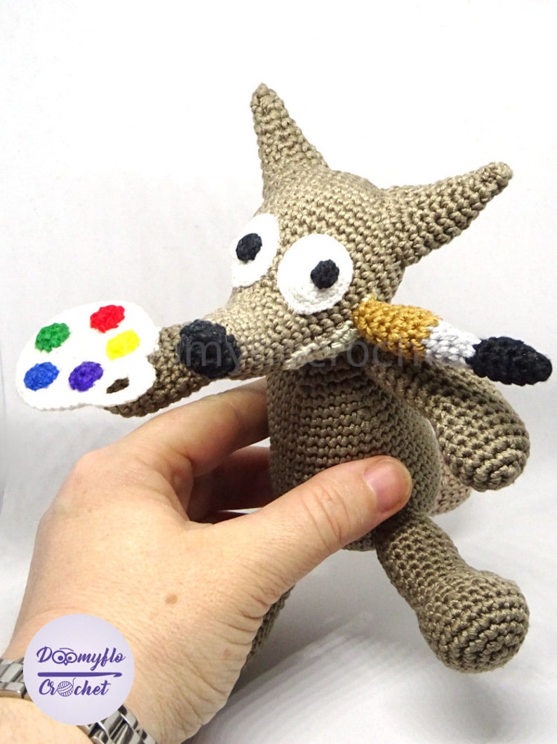 Puede incluir: Un lobo de crochet gris que sostiene una paleta de pintura blanca con puntos de pintura de colores y un pincel en la boca.