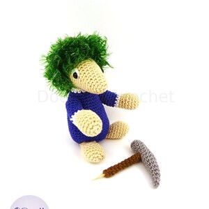 May include: A crocheted green-haired gnome wearing a blue shirt and holding a small grey pickaxe. The gnome is sitting on a white surface.