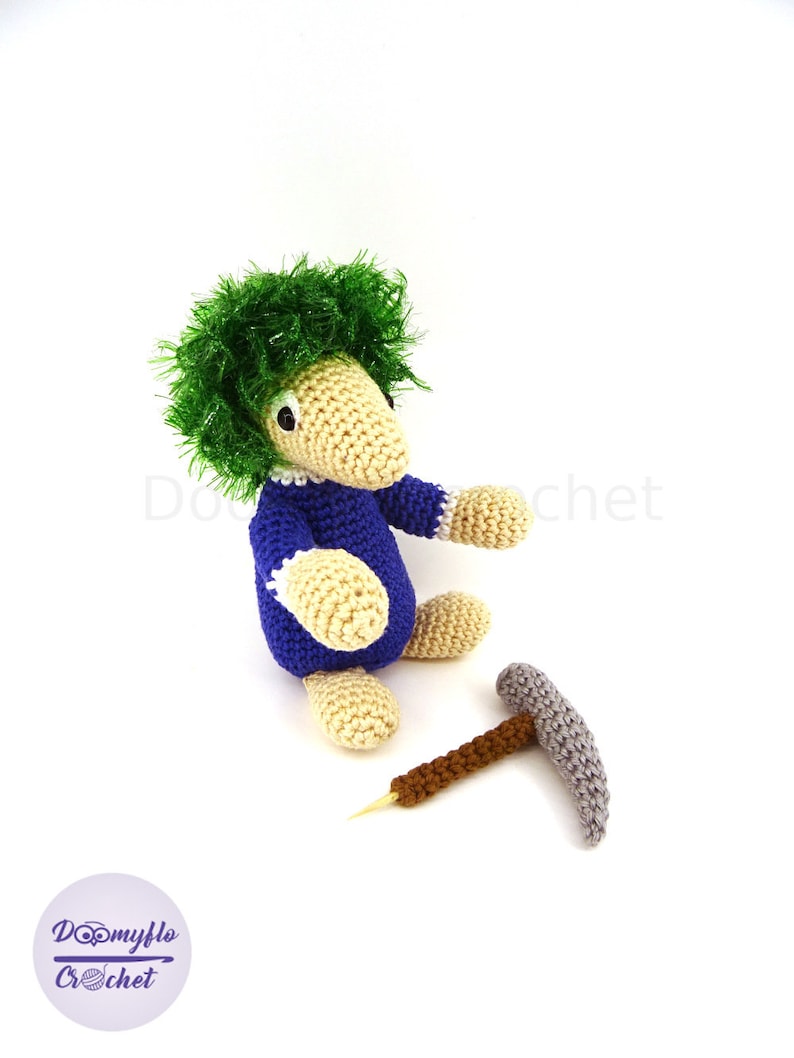 May include: A crocheted green-haired gnome wearing a blue shirt and holding a small grey pickaxe. The gnome is sitting on a white surface.