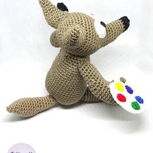 Puede incluir: Un lobo marr&oacute;n de crochet sosteniendo una paleta de pintura blanca con pintura roja, amarilla, azul, verde y naranja.