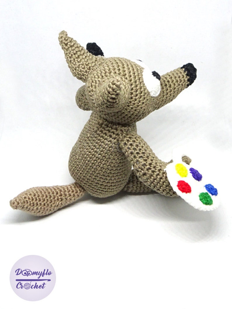 Puede incluir: Un lobo marr&oacute;n de crochet sosteniendo una paleta de pintura blanca con pintura roja, amarilla, azul, verde y naranja.