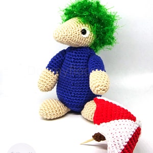 May include: A crocheted toy with green hair, a blue shirt, and beige skin. The toy is holding a red and white crocheted umbrella.