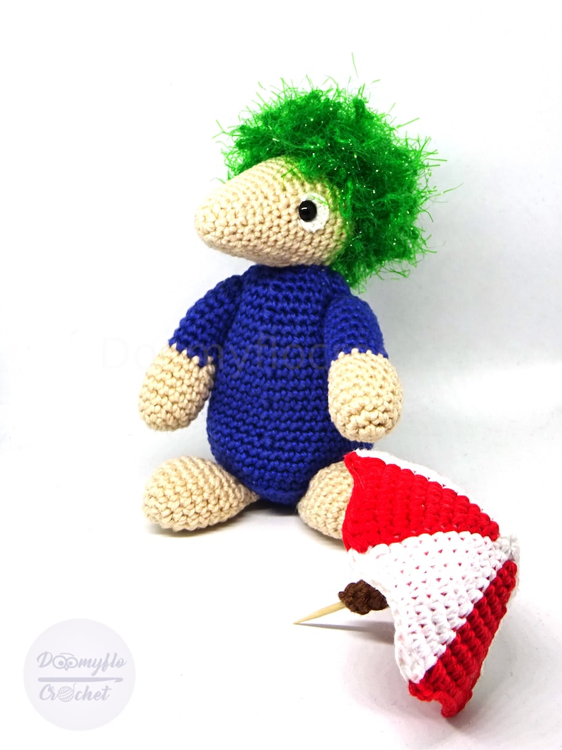 May include: A crocheted toy with green hair, a blue shirt, and beige skin. The toy is holding a red and white crocheted umbrella.