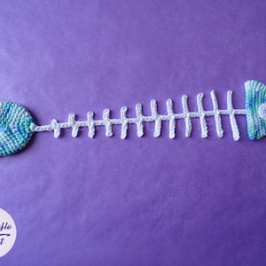 May include: A crocheted fish skeleton with a blue and green tail and head. The skeleton is white and the head has a blue eye.
