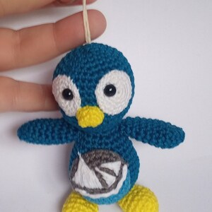 May include: A handmade teal crochet penguin keychain with white accents around the eyes and a yellow beak and feet. The penguin has a small embroidered design on its belly and is attached to a metal keyring.