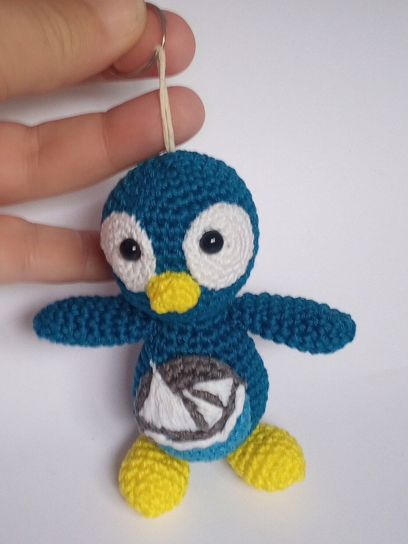 May include: A handmade teal crochet penguin keychain with white accents around the eyes and a yellow beak and feet. The penguin has a small embroidered design on its belly and is attached to a metal keyring.