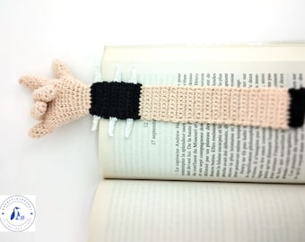 Puede incluir: Un marcapáginas de ganchillo con una mano beige haciendo un gesto de rock and roll, unido a una tira rectangular larga, plana y de color beige. El diseño incluye una sección de puño negro con tres púas blancas a cada lado y una punta negra.
