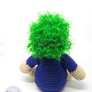 May include: A crocheted plush toy with a blue shirt and green hair. The toy is sitting on a white surface.