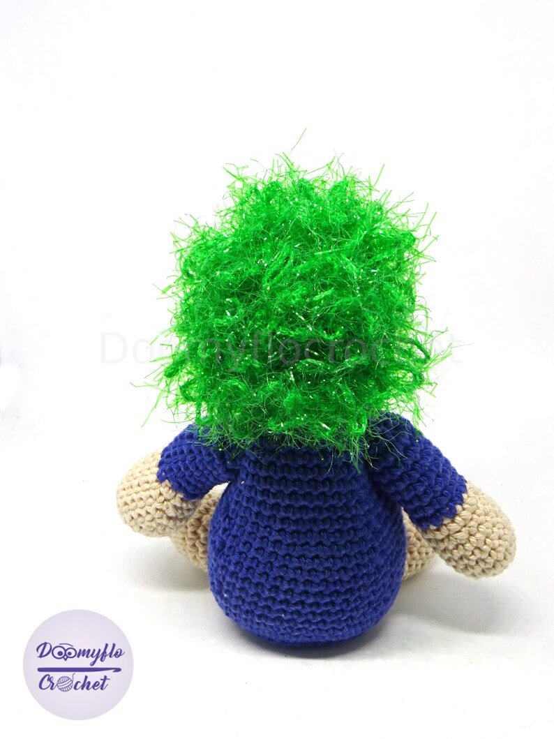 May include: A crocheted plush toy with a blue shirt and green hair. The toy is sitting on a white surface.