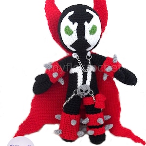 May include: A crocheted plush toy of the superhero Spawn. The toy is black and red with a white face and green eyes. The toy is wearing a red cape and has a chain around its waist. The toy is standing on a white background.