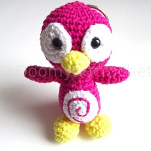May include: A handmade crochet penguin keychain in bright pink, white, and yellow. The penguin has large white eyes with black pupils, a yellow beak and feet, and a white spiral design on its belly. The keychain is a cute, whimsical accessory.