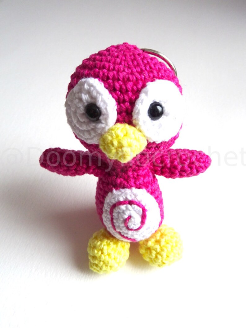 May include: A handmade crochet penguin keychain in bright pink, white, and yellow. The penguin has large white eyes with black pupils, a yellow beak and feet, and a white spiral design on its belly. The keychain is a cute, whimsical accessory.