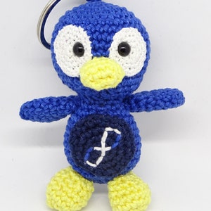 May include: A handmade blue crochet penguin keychain with white eyes, a yellow beak, and yellow feet. The penguin has a dark blue belly with a white "f" design. A silver key ring is attached to the top of the penguin's head.