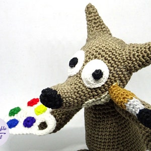 Puede incluir: Un lobo de crochet sosteniendo un pincel y una paleta de pintura blanca con puntos de pintura de colores. El lobo tiene ojos grandes blancos con pupilas negras y una nariz marr&oacute;n.