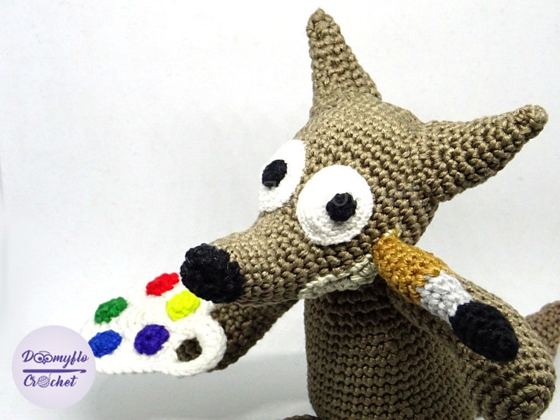 Puede incluir: Un lobo de crochet sosteniendo un pincel y una paleta de pintura blanca con puntos de pintura de colores. El lobo tiene ojos grandes blancos con pupilas negras y una nariz marr&oacute;n.