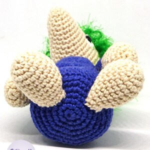 May include: A crocheted toy in the shape of a blue bird with beige wings and a green and white head. The bird is lying on its back with its wings spread out.